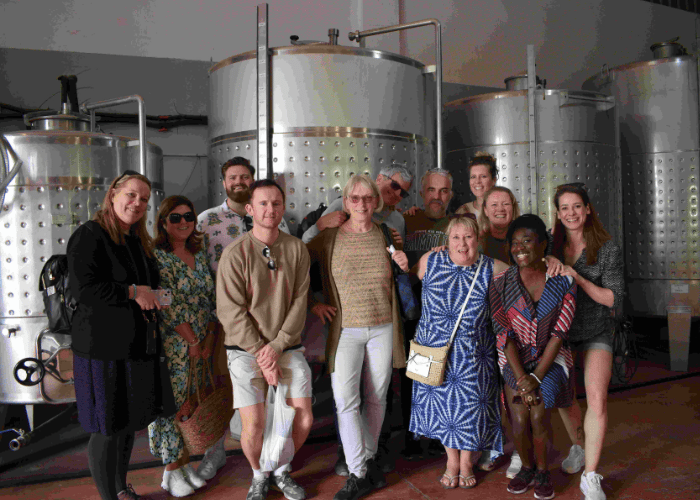 Group of visitors smiling inside a winery in Malaga during a guided wine tour, standing in front of stainless steel fermentation tanks.